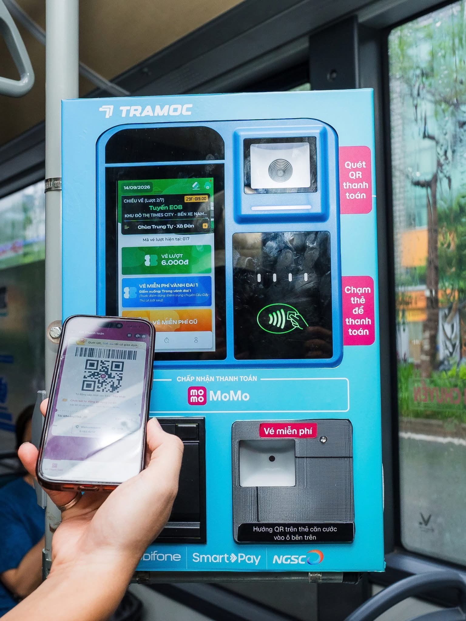Ảnh bàn tay cầm điện thoại có mã QR trước thiết bị thanh toán MoMo màu xanh trong xe buýt.